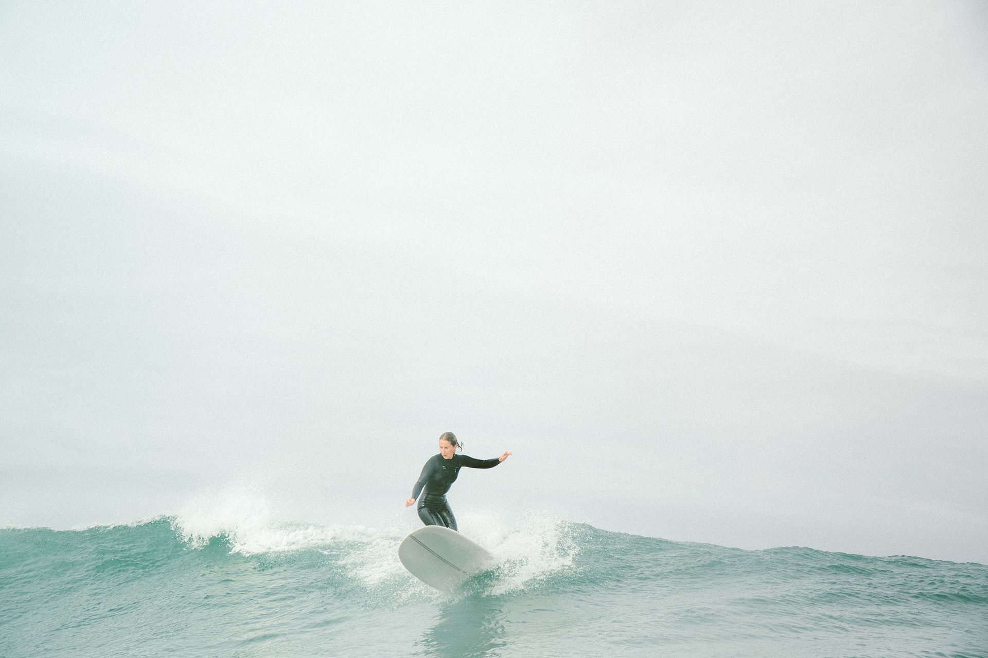 Longboarder in a black wetsuit trimming across a small glassy grey-green wave.