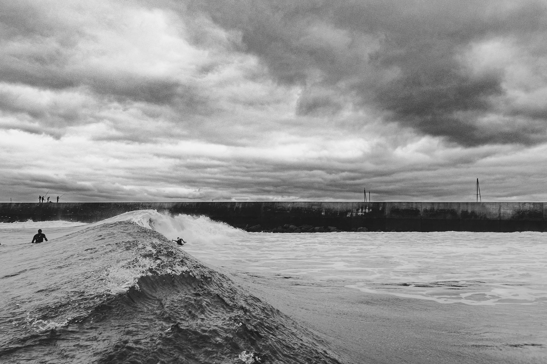 Black and white frame of a bodyboarder speeding along a wave beside the Peniche harbour wall.