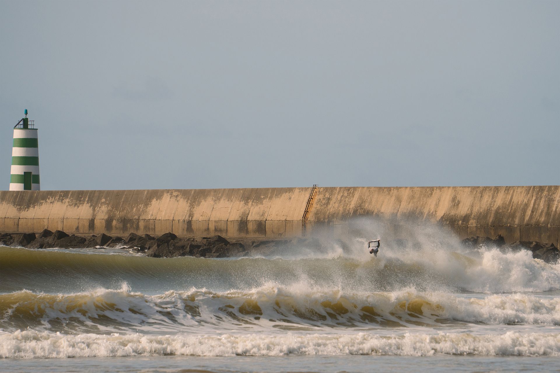 Surfer throwing spray off the lip below the green-striped Peniche lighthouse and breakwater.