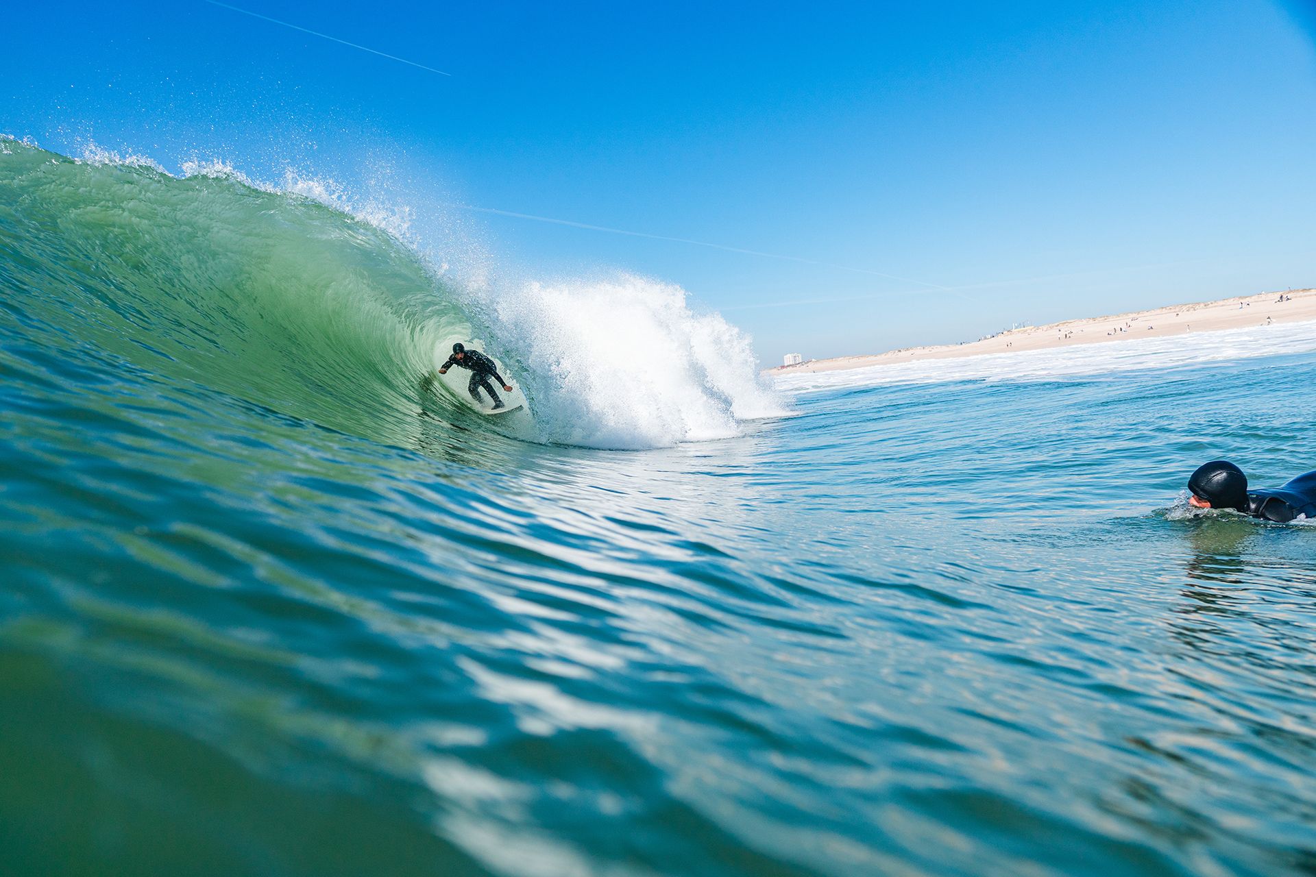 Surfer tucked inside a bright turquoise barrel on a sunny beach-break day as another paddles past.