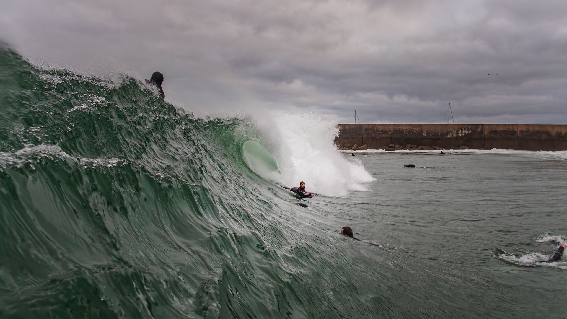 Bodyboarder dropping into a thick green wall as it wraps toward the harbour wall.