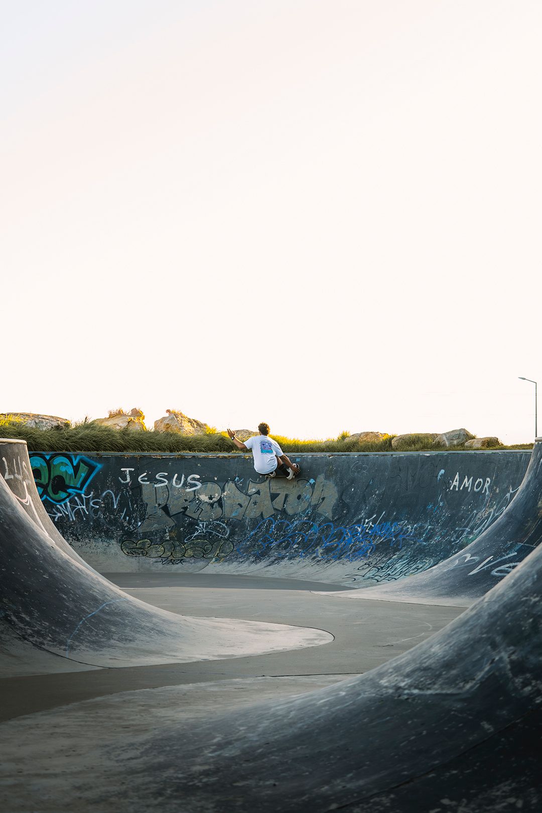 Skater rolling into a graffiti-covered concrete bowl at dusk, the ocean beyond.