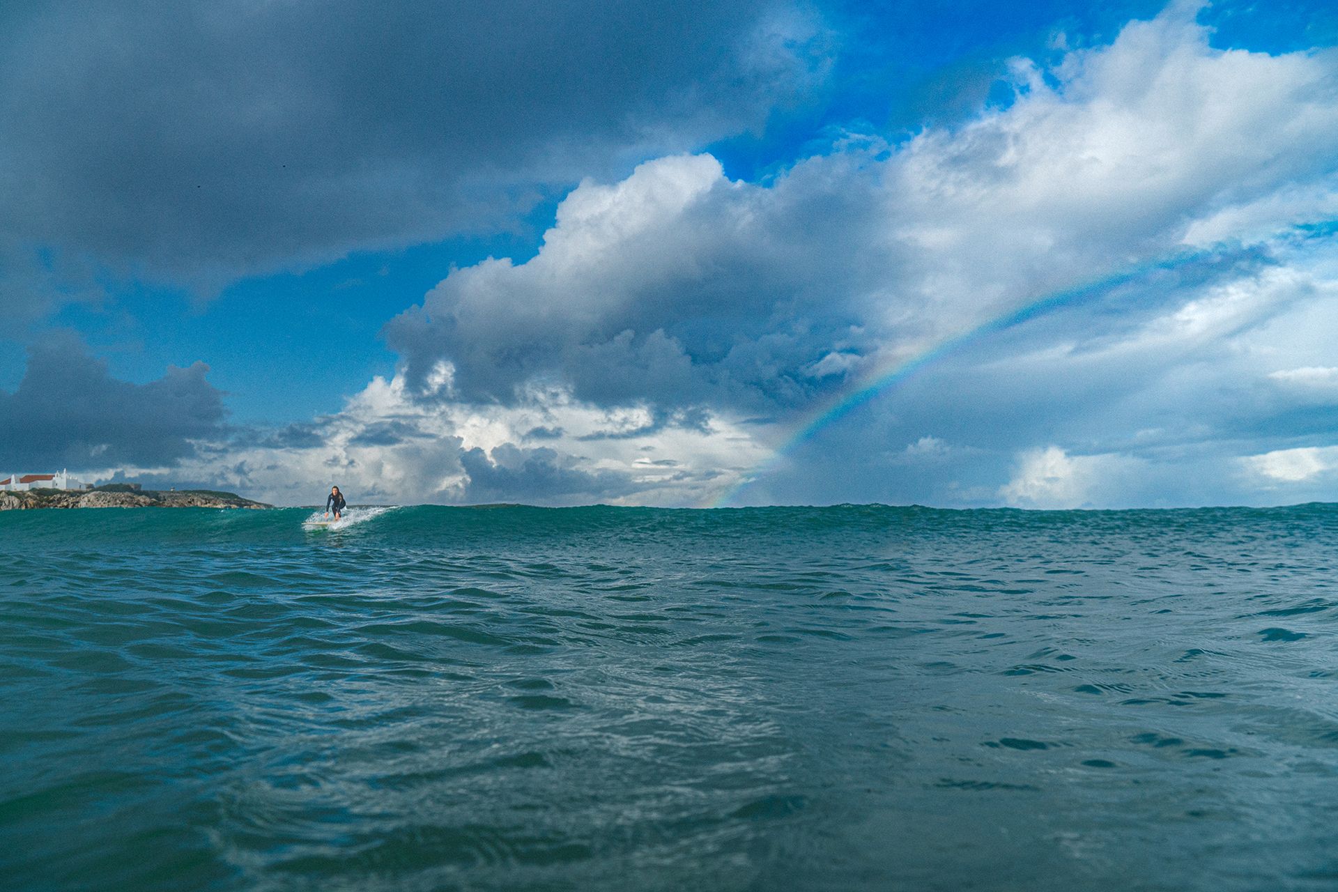 Longboarder gliding over flat water toward a faint rainbow and a distant chapel on the point.