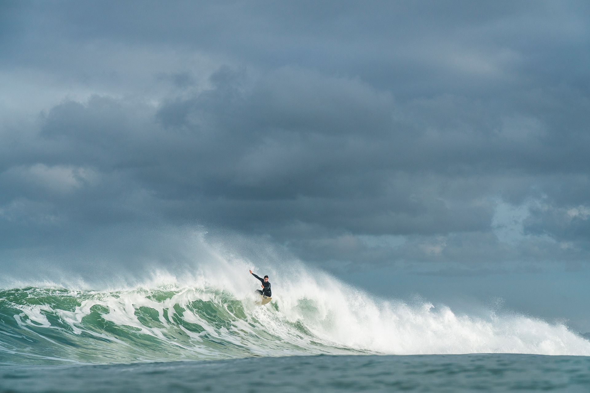 Surfer launching off the lip of a stormy, wind-textured wave under heavy cloud.