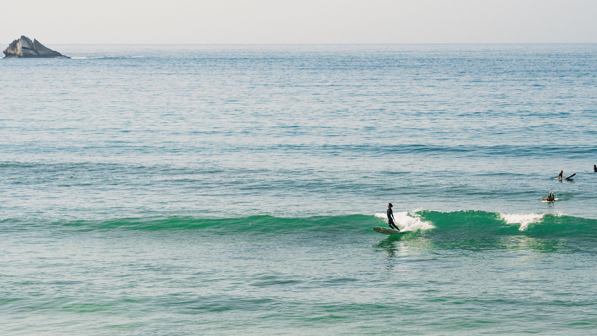 Wide seascape with a small longboarder gliding along a green wave line, a sea stack on the horizon.