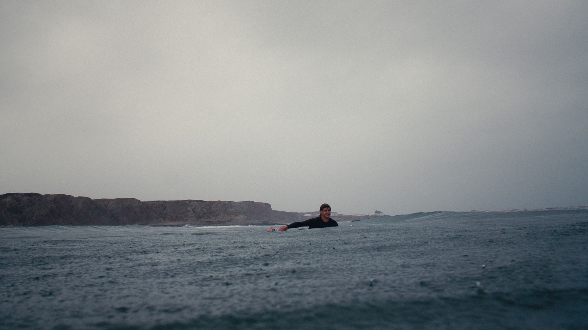 Surfer sitting in a flat grey line-up, smiling, with the Baleal headland behind on an overcast day.