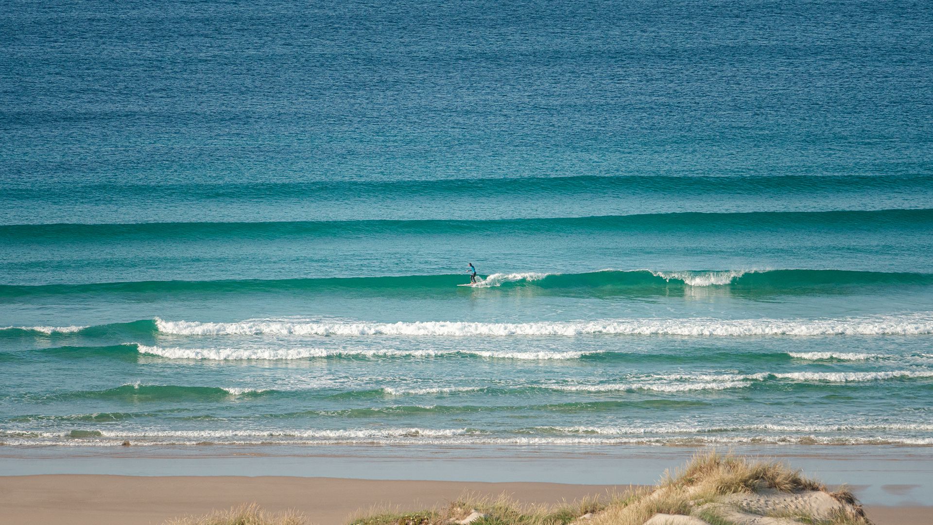 Distant lone surfer walking the nose across a wide expanse of turquoise sandbank lines.