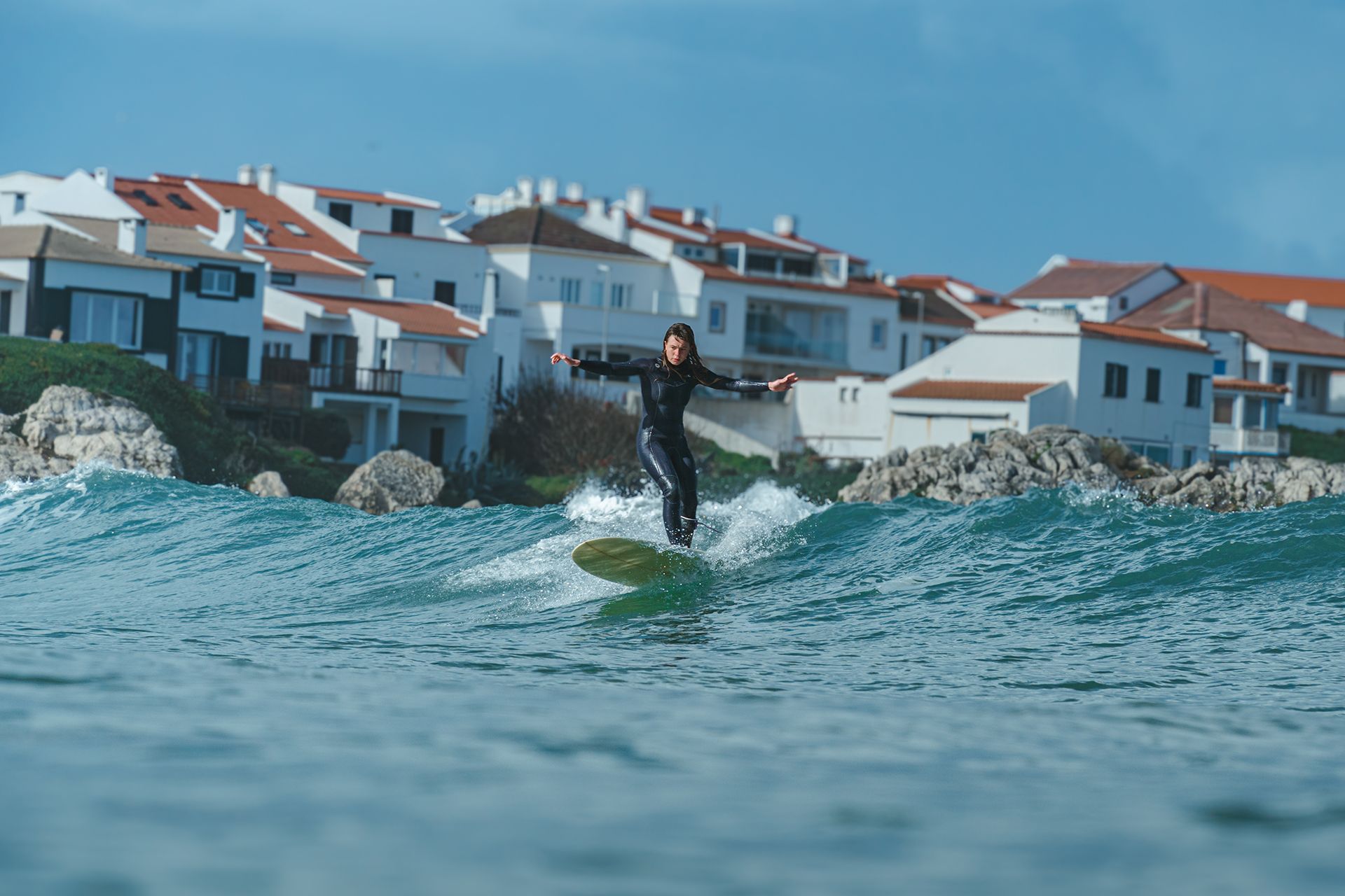Woman noseriding a clean blue wave in front of the white red-roofed houses of Baleal.