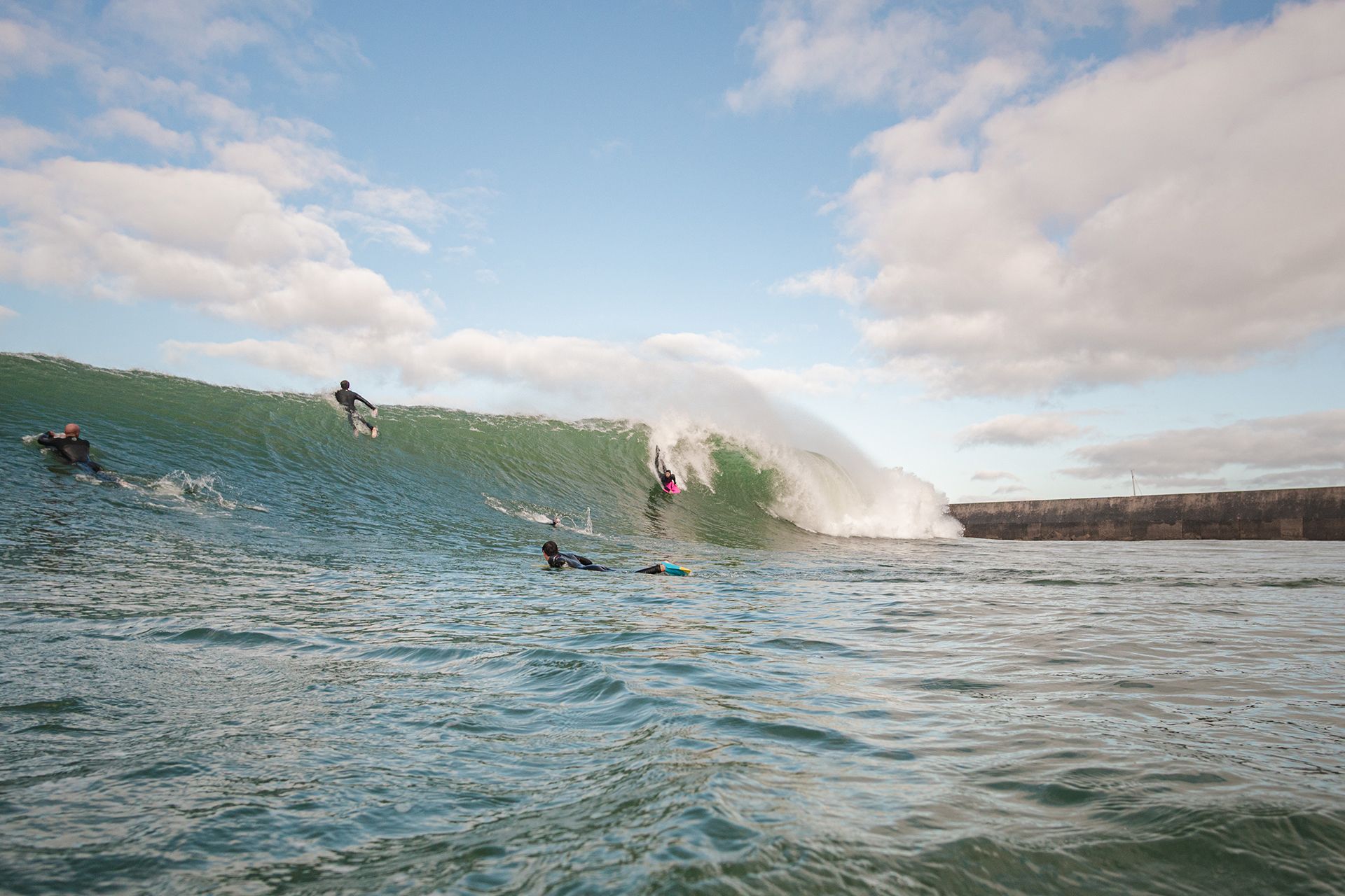 Bodyboarders and a surfer taking a large open-ocean wave beside the breakwater under a blue sky.
