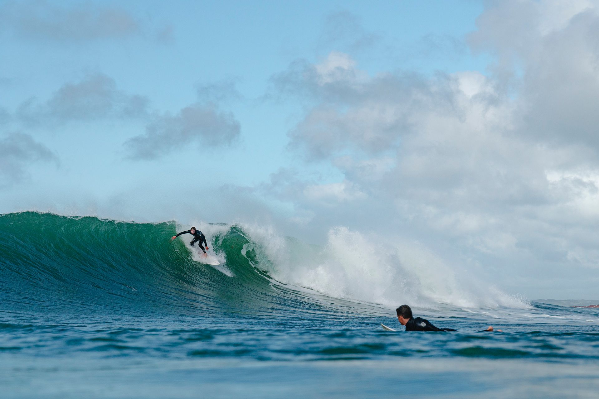 Surfer dropping in at the top of a head-high green wall while another paddles over the shoulder.