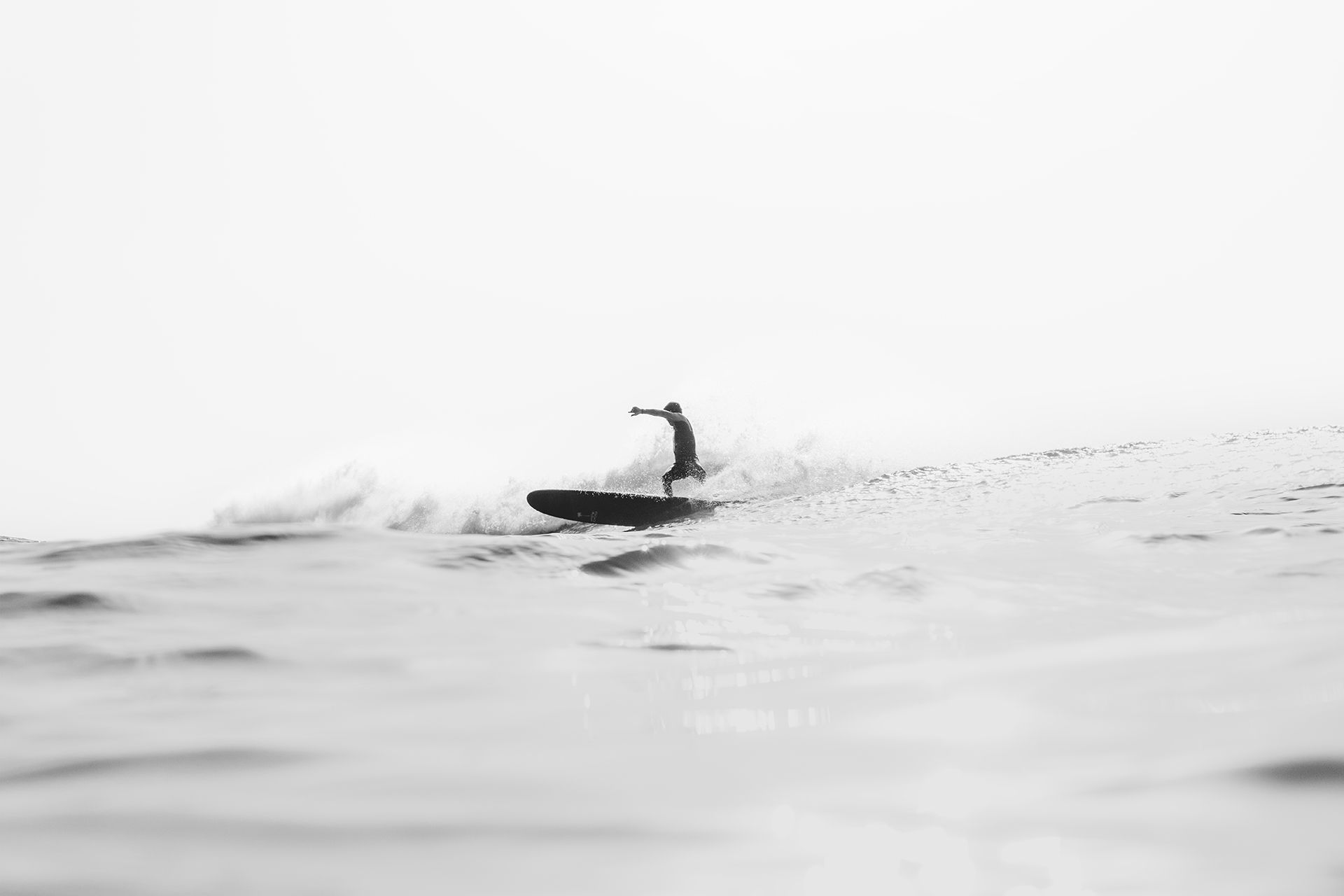 Minimal high-key frame of a surfer stalling on a foamy wave against a white sky.