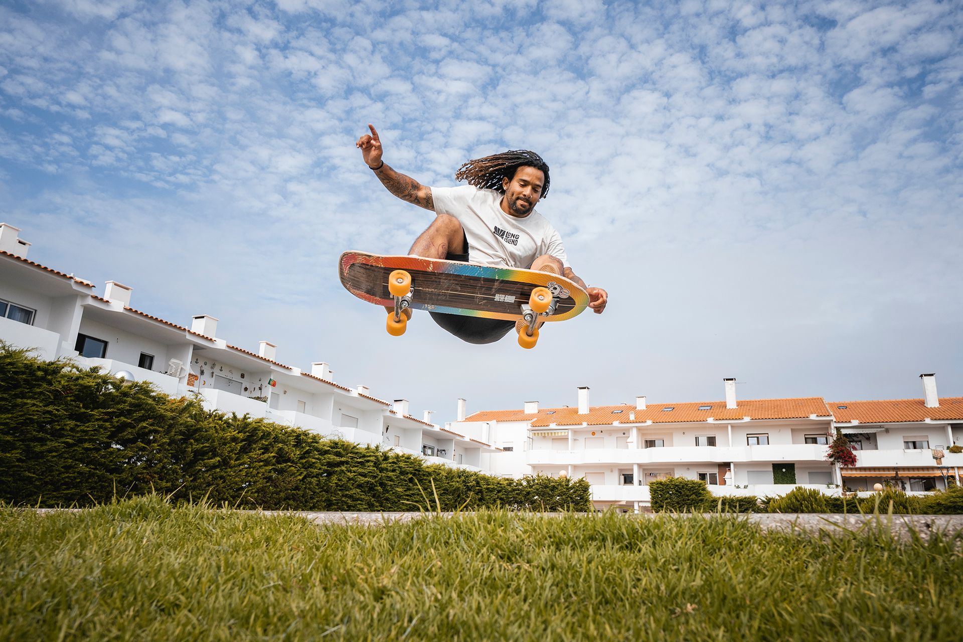 Skater grabbing his board mid-air over a grass verge, white apartment blocks behind.