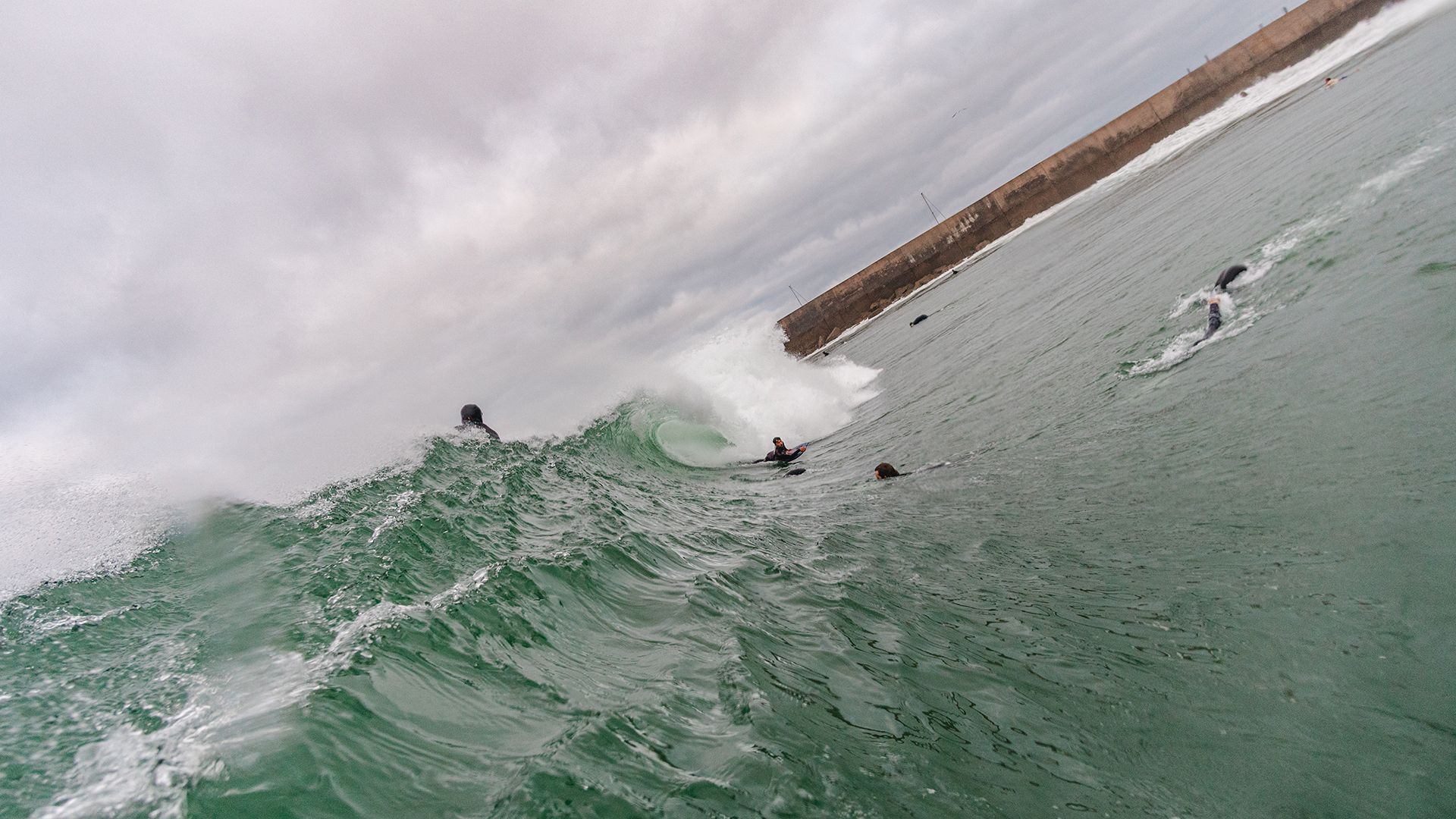 Wide tilted frame of the harbour-wall wave with bodyboarders and swimmers scattered across it.